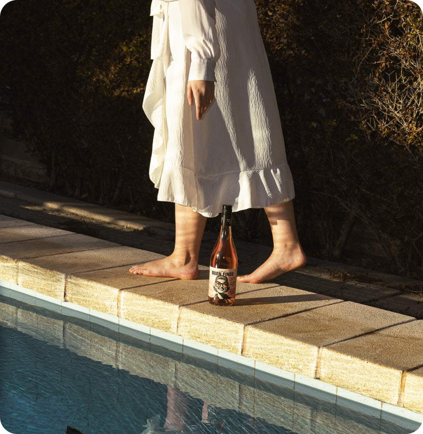Una persona descalza con un vestido blanco camina por el borde de una piscina junto a una botella de Buen Finde